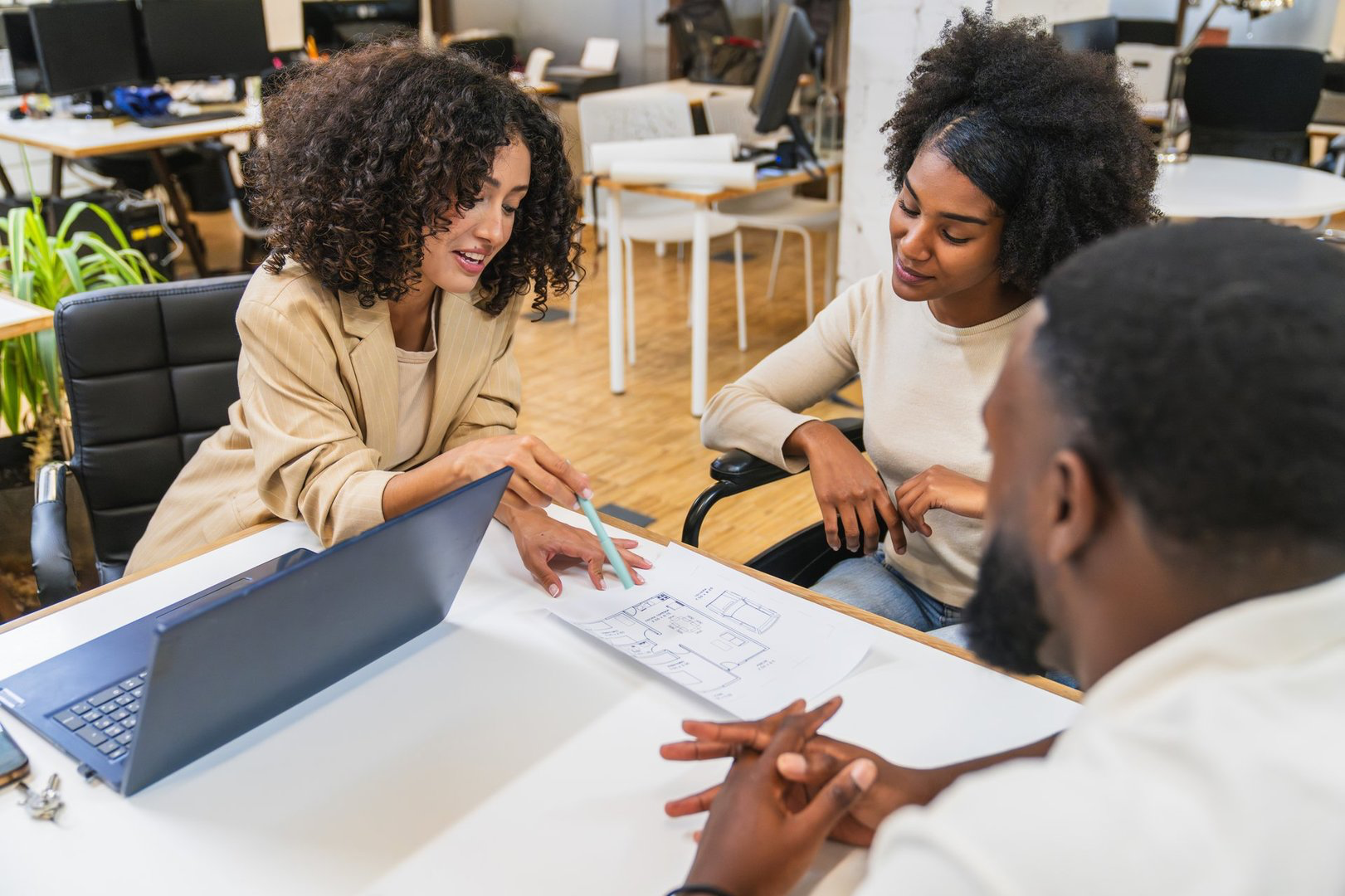 Architect explaining a project to a couple, using a laptop and a blueprint, in a modern coworking space